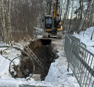 Grävd ränna med nedlagda ledningar, omgiven av jordhögar och grävmaskin i bakgrunden.