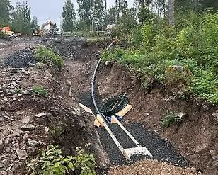 Grävd ränna med nedlagda ledningar, omgiven av jordhögar och grävmaskin i bakgrunden.